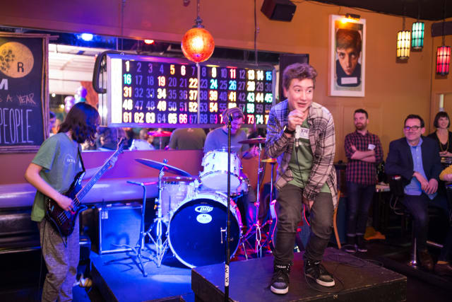 Youth performing on stage at Love Bomb 2017 event, with a band setup featuring guitar and drums, vibrant lighting, and an engaged audience.